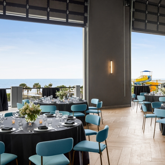 Seaside Vista ballroom above Black Drum Brewing set up in rounds for a banquet event. The garage doors are open with a direct view of the ocean and the terrace has cocktail tables set up.