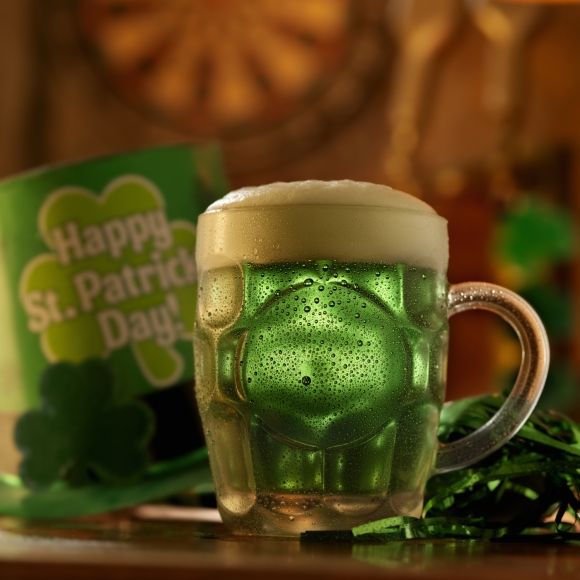 A beer mug full of Green Beer with a sign in the background that says Happy St. Patrick's Day!