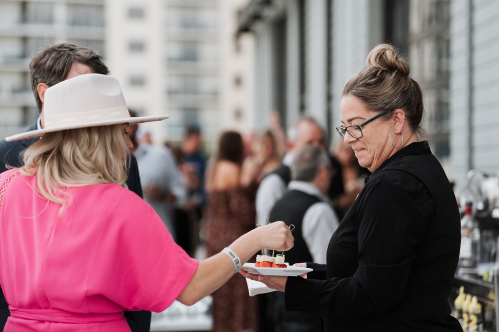 Women in a large white hat taking watermelon and mozzarella passed hor d'oeuvres from a server.