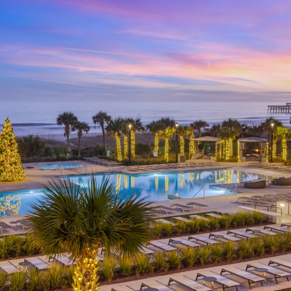 A pool deck lit with christmas lights on all the surrounding palm trees and a 20 foot lit christms tree in the middle of the pool deck. Lounge chair are surround the entire pool