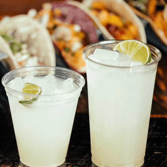 Two glasses with a regular margarita in the forefront of the image. Blurred in the background is a tray of six various soft shell tacos